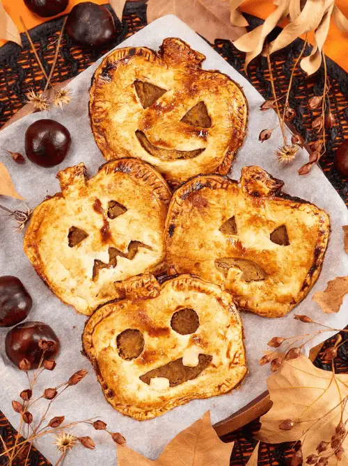 Mini pumpkin hand pies with spooky jack-o’-lantern faces, golden-brown crust, and a flaky texture—festive and fun Halloween treats surrounded by autumn decor.