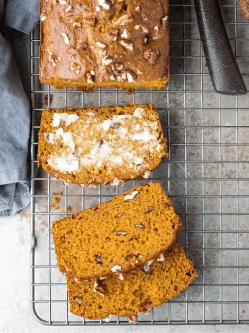 Sliced pumpkin bread with pecans cooling on a wire rack, one slice topped with melted butter — a flavorful example of what to do with leftover pumpkin puree and a nutty twist on easy pumpkin bread recipes.