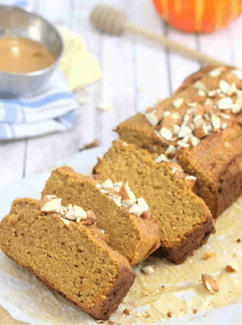 Sliced loaf of pumpkin bread topped with chopped almonds, set on parchment paper with honey and a small bowl in the background.