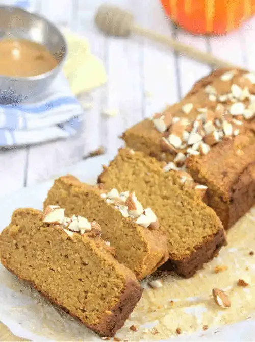 Sliced loaf of pumpkin bread topped with chopped almonds, set on parchment paper with honey and a small bowl in the background.