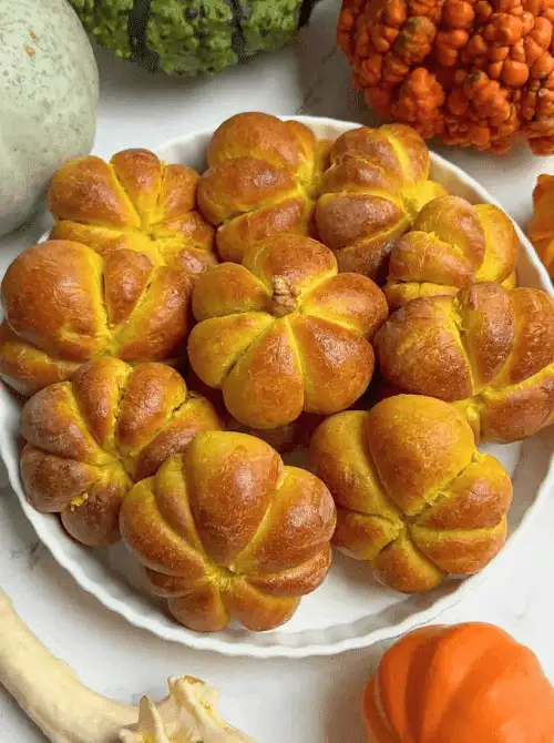 Golden pumpkin-shaped dinner rolls arranged on a white plate, surrounded by fall gourds—festive and budget-friendly make-ahead Halloween party food idea.