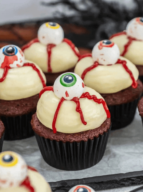 Red velvet cupcakes topped with white frosting, candy eyeballs, and red gel “blood” drizzle—creepy and bold make-ahead Halloween party food for horror-themed dessert tables.