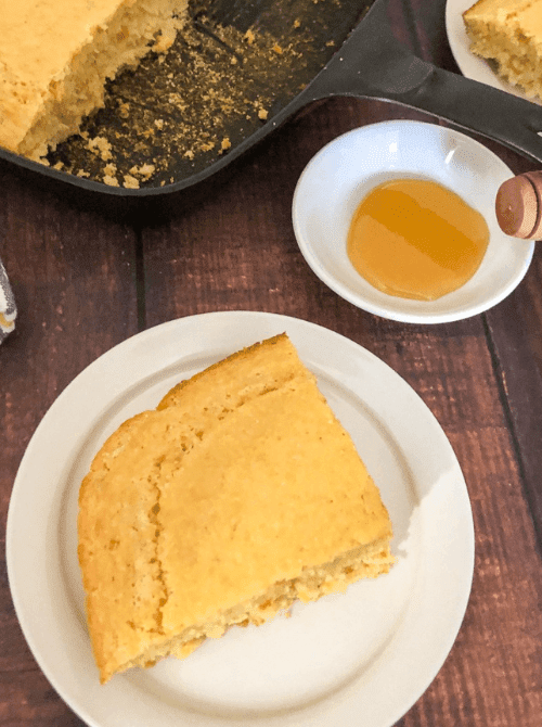 Slice of homemade sweet corn bread served on a white plate with honey, a warm and budget-friendly Thanksgiving side dish.