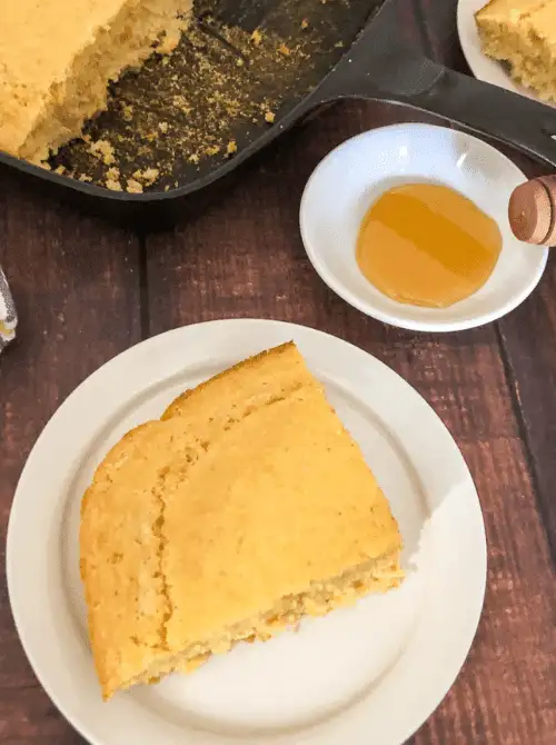Slice of homemade sweet corn bread served on a white plate with honey, a warm and budget-friendly Thanksgiving side dish.