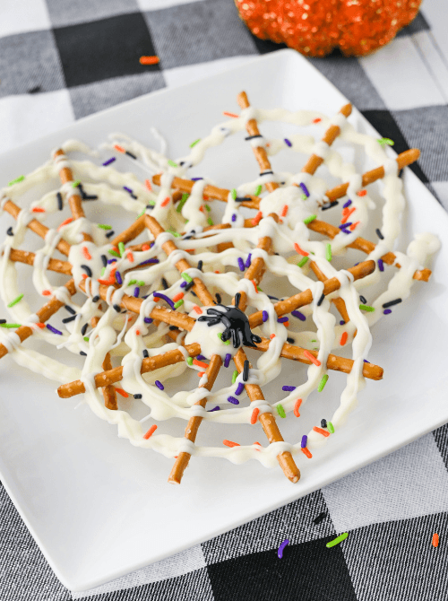 Plate of spider web pretzels drizzled with white chocolate and topped with colorful Halloween sprinkles and plastic spiders — a fun and festive option for No-Bake Halloween Treats.