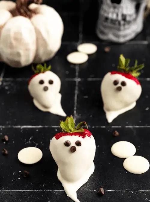 No-Bake Halloween Treats featuring white chocolate-dipped strawberries decorated to look like ghosts with mini chocolate chip eyes, set against a spooky party backdrop.
