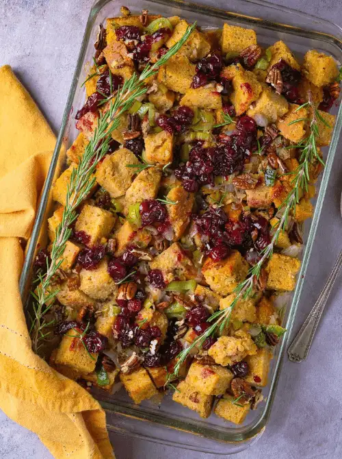 Glass baking dish of vegan cornbread stuffing topped with cranberries, pecans, and fresh rosemary, a colorful budget-friendly Thanksgiving side.