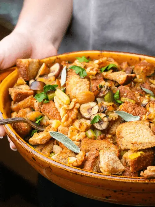 Person holding a rustic baking dish filled with bread stuffing made with celery, grapes, walnuts, and fresh herbs, a unique budget-friendly Thanksgiving side.