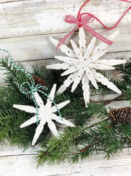 Dollar Store Christmas DIY decor snowflake ornaments made from painted clothespins, decorated with twine and ribbon, displayed on pine greenery with pinecones.
