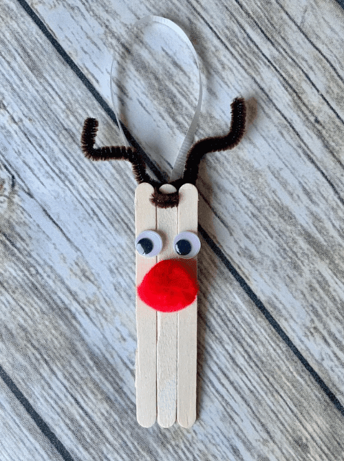 Dollar Store Christmas DIY decor reindeer ornament made from popsicle sticks with googly eyes, red pom-pom nose, and pipe cleaner antlers, tied with a ribbon for hanging.