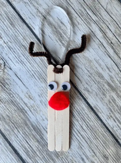 Dollar Store Christmas DIY decor reindeer ornament made from popsicle sticks with googly eyes, red pom-pom nose, and pipe cleaner antlers, tied with a ribbon for hanging.