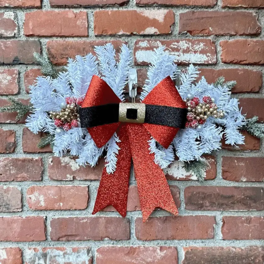 Dollar Store Christmas DIY decor featuring a snowy white wreath with a large red glitter bow and festive accents, hanging on a brick wall.