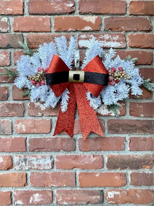 Dollar Store Christmas DIY decor featuring a snowy white wreath with a large red glitter bow and festive accents, hanging on a brick wall.