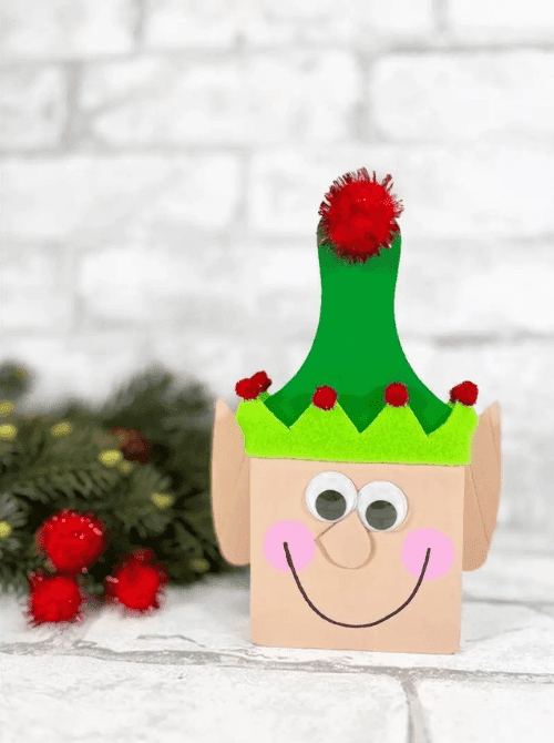 Dollar Store Christmas DIY decor elf craft made from a small box with felt hat, pom-poms, googly eyes, and painted smile, displayed with holiday greenery.