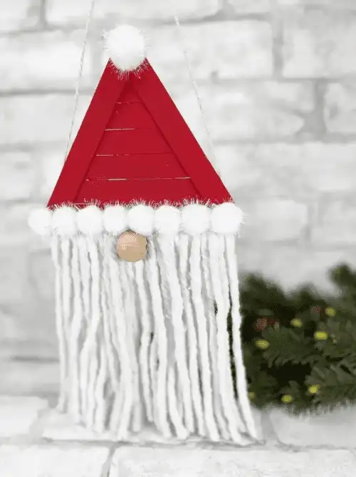 Dollar Store Christmas DIY decor Santa gnome ornament made with a red triangle hat, pom-poms, yarn beard, and wooden nose, hanging against a white brick background.