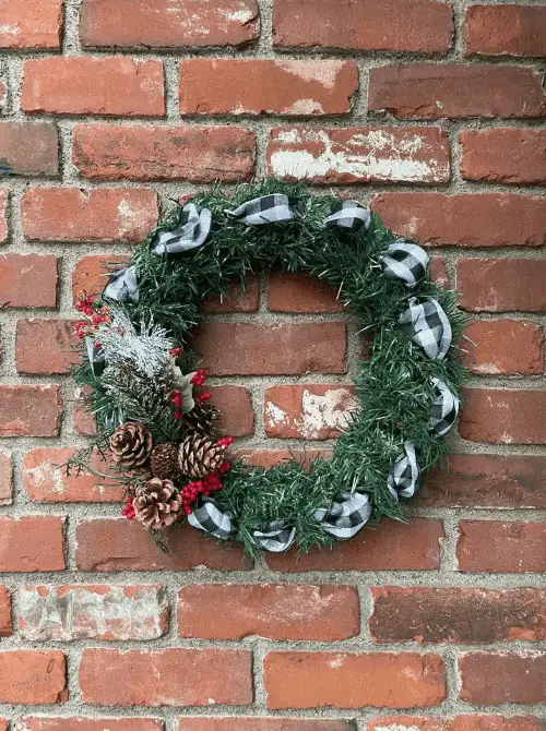 Dollar Store Christmas DIY decor wreath with black and white plaid ribbon, pinecones, red berries, and frosted greenery on a brick wall.