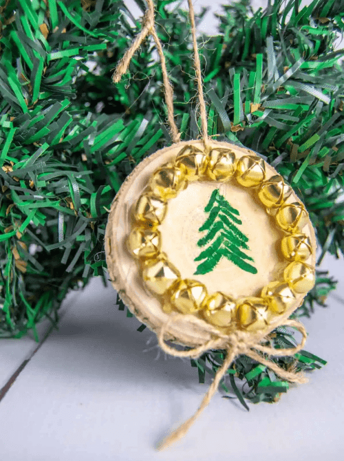 Dollar Store Christmas DIY decor wood slice ornament painted with a green Christmas tree and decorated with gold jingle bells, tied with twine and displayed on a tree.