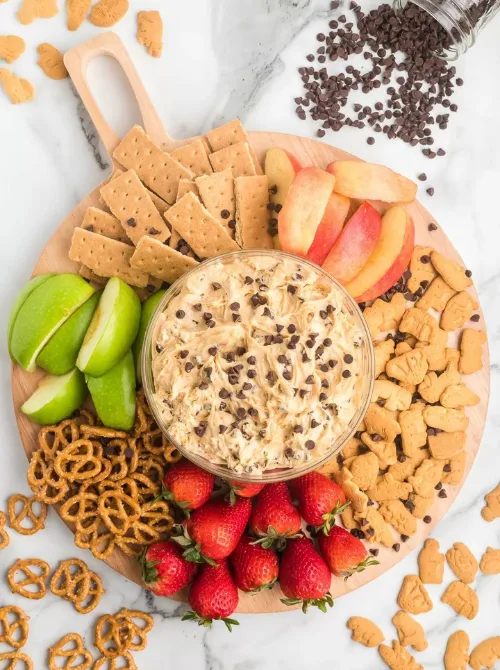 A round wooden platter filled with chocolate chip dessert dip surrounded by graham crackers, apple slices, pretzels, strawberries, and animal crackers — a fun and budget-friendly idea for party dips for the holidays. This easy dessert dip makes a festive centerpiece for any holiday dessert table or family gathering.