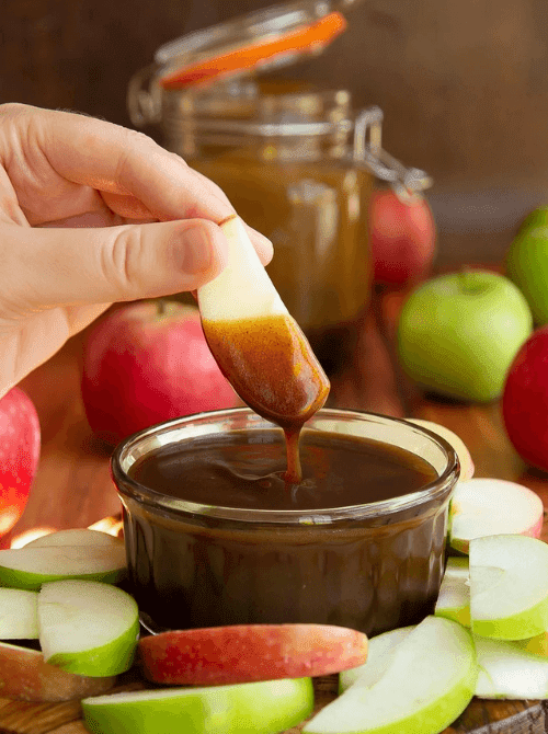 A hand dips a fresh apple slice into a rich caramel easy dessert dip, surrounded by red and green apples on a rustic wooden table. Perfect for party dips for the holidays, this cozy fall-inspired treat is ideal for festive gatherings and sweet snack boards.
