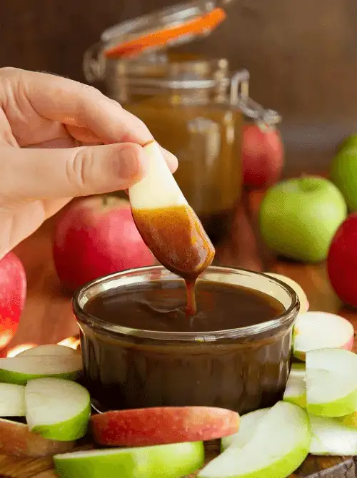 A hand dips a fresh apple slice into a rich caramel easy dessert dip, surrounded by red and green apples on a rustic wooden table. Perfect for party dips for the holidays, this cozy fall-inspired treat is ideal for festive gatherings and sweet snack boards.