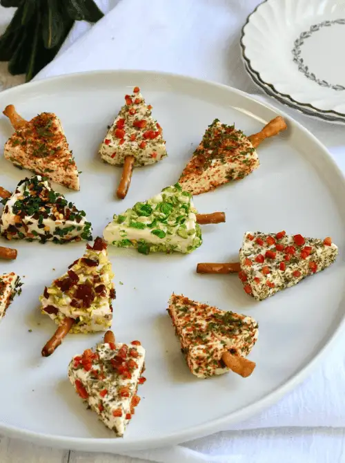 Festive tree-shaped cheese appetizer bites with pretzel sticks arranged on a white plate, topped with colorful herbs and peppers — a fun example of make-ahead Christmas appetizers and easy Christmas party food ideas.