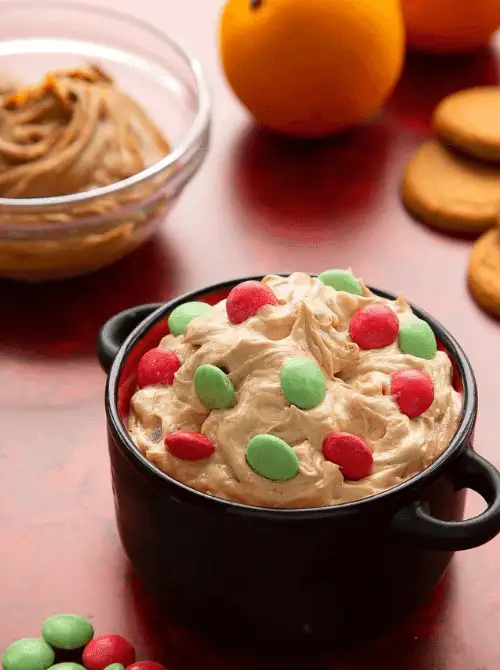 A festive bowl of easy dessert dip swirled with creamy brown sugar and topped with red and green candies, perfect for party dips for the holidays. The dip is served in a black bowl with cookies and oranges in the background on a red surface, creating a cheerful Christmas party vibe.
