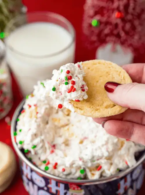A festive bowl of fluffy easy dessert dip topped with red, green, and white sprinkles, with a hand dipping a cookie into the creamy mixture. Perfect for party dips for the holidays, this sweet Christmas treat pairs beautifully with milk and sugar cookies for a cheerful seasonal spread.