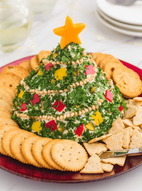 Christmas tree–shaped cheese ball coated in herbs and decorated with peppers, served with crackers, a festive centerpiece for make-ahead Christmas appetizers and fun Christmas party food ideas.