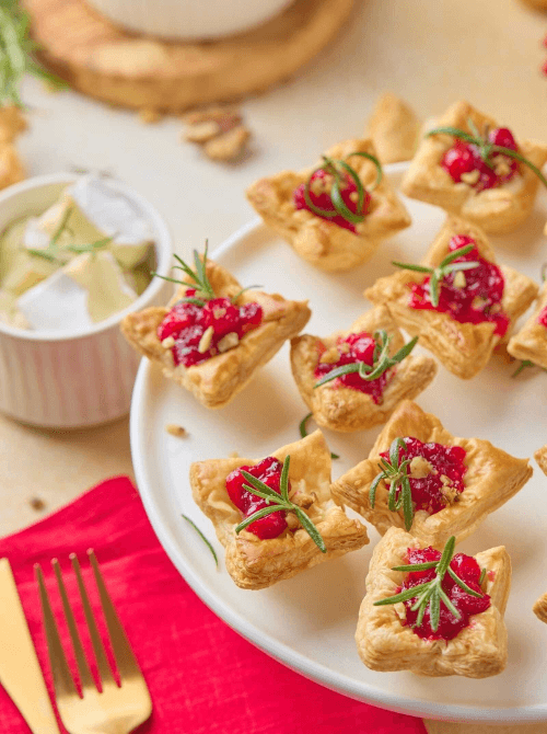 Mini puff pastry bites filled with cranberry and topped with rosemary on a white plate, a festive and easy option for make-ahead Christmas appetizers and Christmas party food ideas.