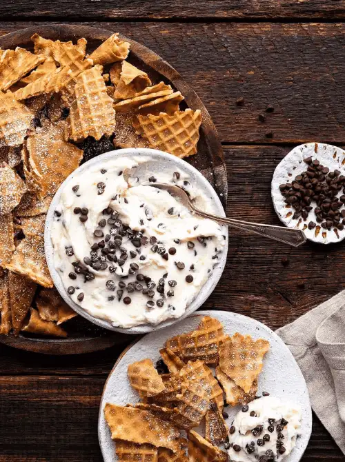 A creamy cannoli-inspired easy dessert dip topped with mini chocolate chips, served with crisp waffle cone pieces on rustic plates. Perfect for party dips for the holidays, this indulgent sweet dip combines a festive look with a cozy, homemade charm ideal for sharing at gatherings.
