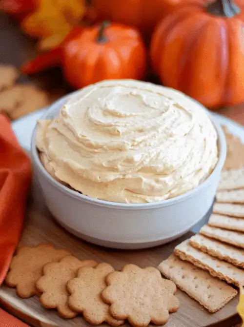 A fluffy pumpkin spice easy dessert dip served in a white bowl, surrounded by gingersnaps and graham crackers on a wooden tray. Small pumpkins and fall decor in the background create a festive, cozy look — perfect for party dips for the holidays or autumn gatherings.