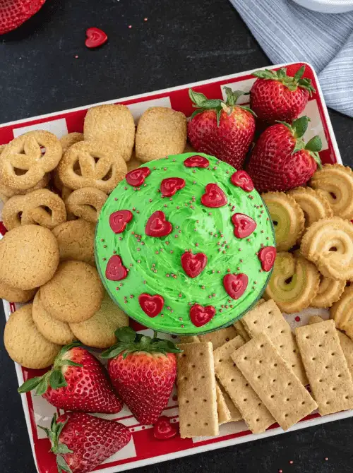 A festive green easy dessert dip topped with red heart-shaped decorations, surrounded by cookies, graham crackers, and fresh strawberries on a red and white tray. Perfect for party dips for the holidays, this colorful treat adds a fun and whimsical touch to any Christmas dessert spread.