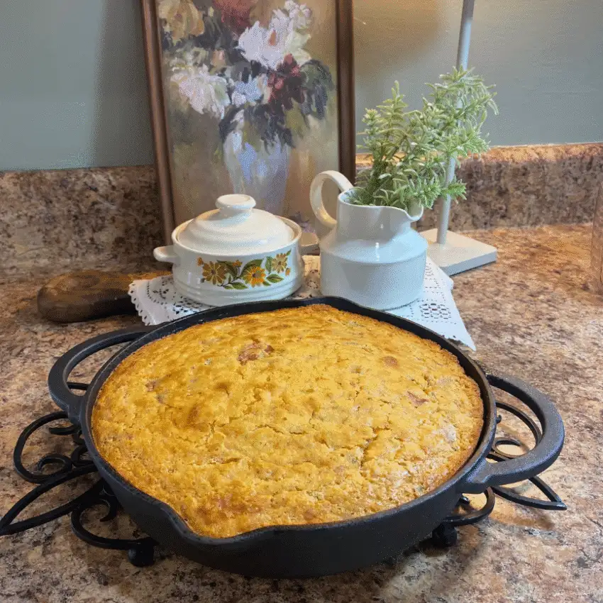 Freshly baked Jiffy Cornbread Casserole with Tomatoes and Okra in a cast-iron skillet, golden brown on top, displayed on a cozy kitchen counter with vintage décor.