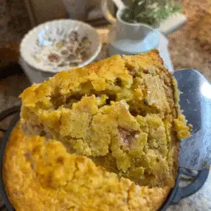 A warm slice of Jiffy Cornbread Casserole with Tomatoes and Okra lifted from a cast-iron skillet, showing its moist, golden texture with bits of corn and tomato in a cozy kitchen setting.