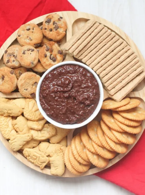 Chocolate hummus served in a white bowl surrounded by cookies, crackers, and graham wafers on a wooden platter — a creative, budget-friendly idea for party dips for the holidays. This easy dessert dip combines sweet and savory flavors, perfect for festive gatherings or family celebrations.