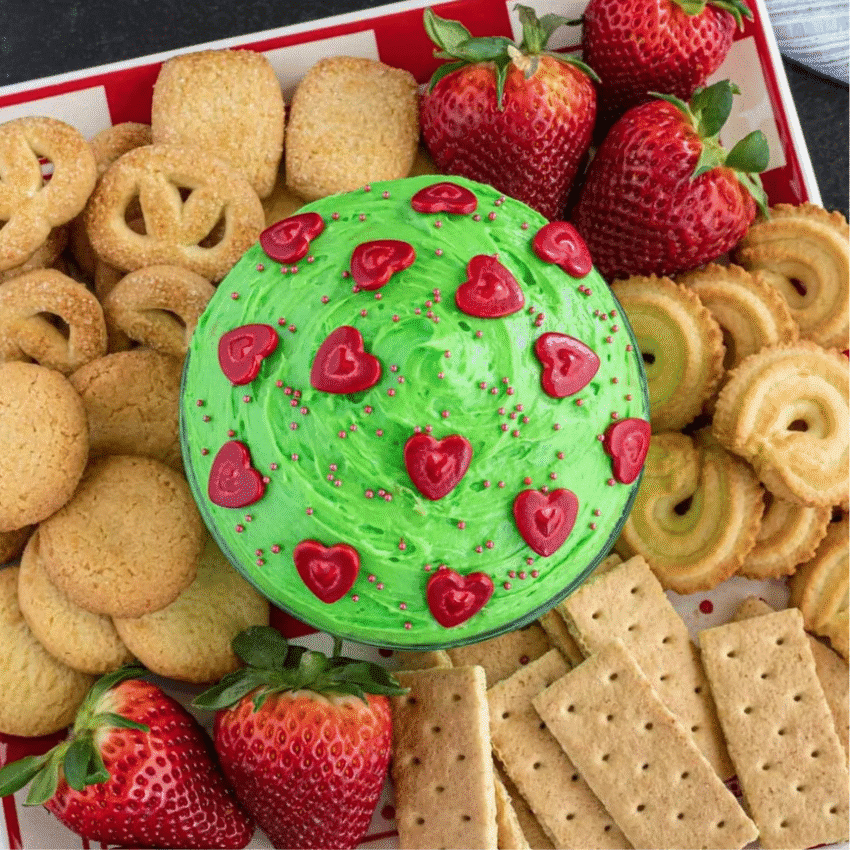 A festive green easy dessert dip topped with red heart-shaped decorations, surrounded by cookies, graham crackers, and fresh strawberries on a red and white tray. Perfect for party dips for the holidays, this colorful treat adds a fun and whimsical touch to any Christmas dessert spread.