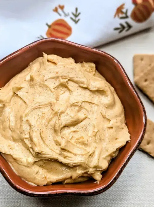 A creamy bowl of pumpkin-flavored easy dessert dip with warm fall spices, served in a rustic brown dish. Perfect for party dips for the holidays, this festive treat is surrounded by graham crackers and autumn-themed napkins for a cozy seasonal presentation.