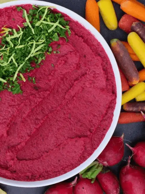 Bright red roasted beet hummus topped with herbs and lemon zest, served with fresh vegetables, a colorful choice for Party Dips for the Holidays with Global Twists.