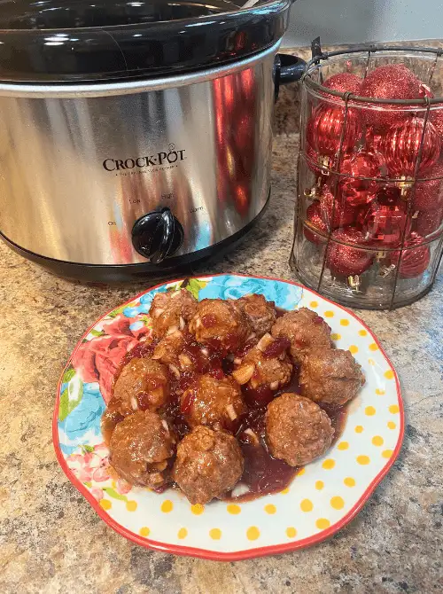 Sweet chili cranberry crockpot meatballs served on a floral plate with a Crock-Pot slow cooker and red holiday ornaments in the background, shown as an easy Christmas appetizer for a crowd.