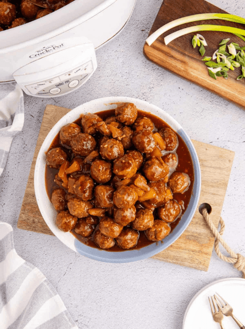 Sweet and sour meatballs served in a bowl with pineapple pieces beside a slow cooker, a budget-friendly favorite for make-ahead Christmas appetizers and holiday Christmas party food ideas.