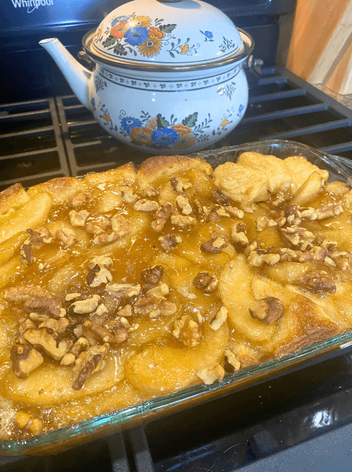 Glass baking dish filled with homemade apple monkey bread topped with apple slices, golden cinnamon glaze, and chopped walnuts, shown fresh from the oven next to a floral teapot.