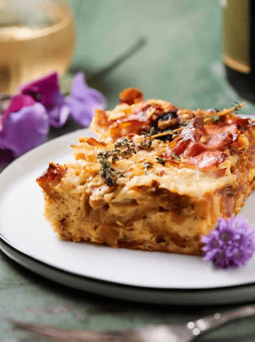 Slice of French onion croissant casserole with herbs on a white plate, an elegant and savory option for Christmas morning breakfast featured in casseroles and quiche recipes.