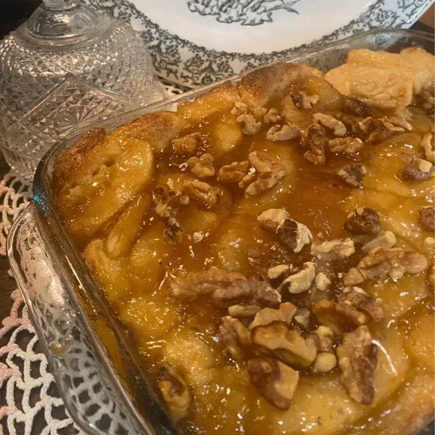 A close-up of a warm apple monkey bread baked in a square glass dish, topped with glossy caramel sauce and toasted walnuts. The golden biscuits and tender apple filling create a gooey, pull-apart texture, set against a vintage-style table setting with a crystal jar and decorative plate in the background.