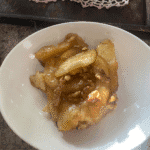 A close-up of a warm, gooey slice of homemade monkey bread made with canned biscuits, coated in golden caramel sauce and topped with tender apple slices and crunchy walnuts, served in a white bowl.
