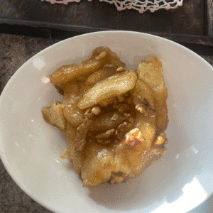 A close-up of a warm, gooey slice of homemade monkey bread made with canned biscuits, coated in golden caramel sauce and topped with tender apple slices and crunchy walnuts, served in a white bowl.
