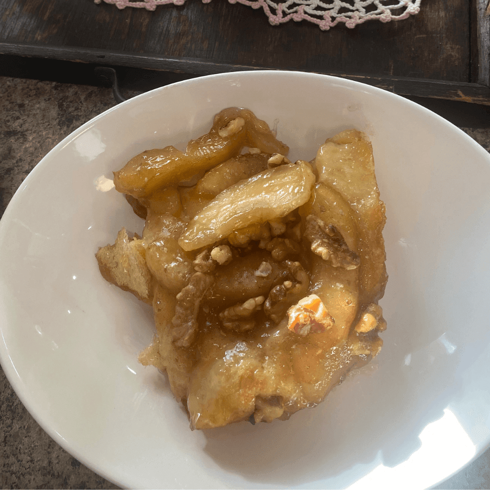 A close-up of a warm, gooey slice of homemade monkey bread made with canned biscuits, coated in golden caramel sauce and topped with tender apple slices and crunchy walnuts, served in a white bowl.