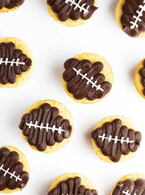 Football-shaped cupcakes topped with chocolate frosting and white icing laces arranged on a white background, perfect Super Bowl party food for a football-shaped dessert table.