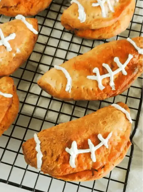 Golden baked buffalo chicken hand pies shaped like footballs with white icing laces cooling on a wire rack, perfect football-shaped Super Bowl party food.