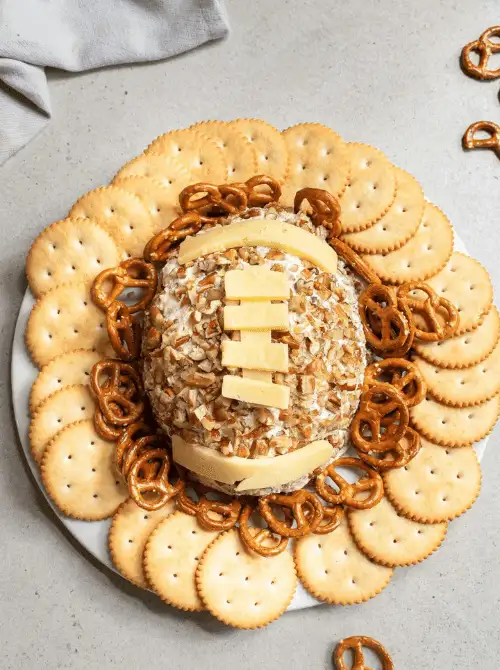 Football-shaped cheese ball coated in chopped nuts, topped with cheese “laces,” and surrounded by crackers and pretzels on a white platter, perfect Super Bowl party food centerpiece.