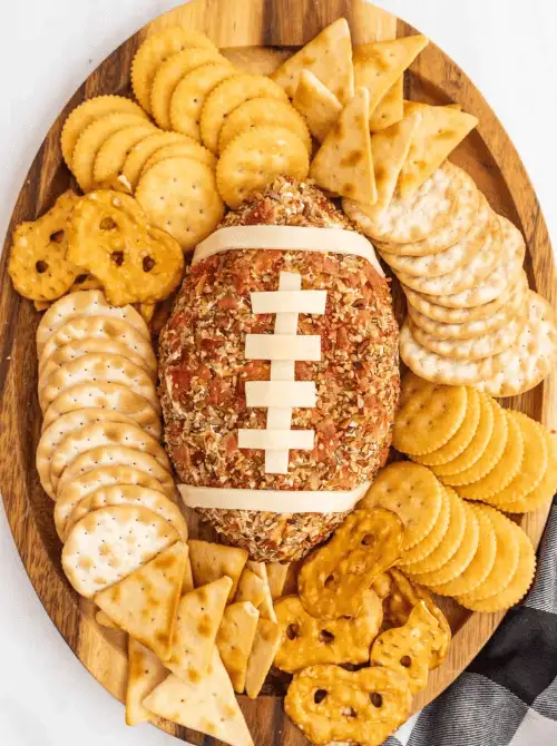 Football-shaped cheese ball covered in seasoning and topped with cheese laces, surrounded by crackers and pretzels on a wooden board, styled as Super Bowl party food for a football-shaped buffet table.